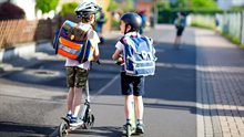 Two school kid boys in safety helmet riding with scooter in the city with backpack on sunny day. Happy children in colorful clothes biking on way to school. Safe way for kids outdoors to school.